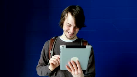 Student-man-uses-a-tablet-for-video-communication-and-chatting-with-friends-on-a-blue-wall-background