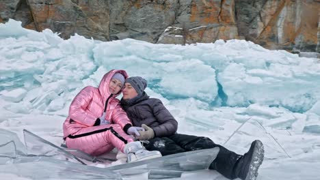 Young-couple-has-fun-during-winter-walk-against-background-of-ice-of-frozen-lake.-Lovers-sit-on-ice,-kiss-and-hug.-Young-happy-people-stroll-through-snow-covered-ice.-Honeymoon.-Love-story.