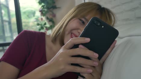 Asian-woman-playing-smartphone-while-lying-on-home-sofa-in-her-living-room.-Happy-female-use-phone-for-texting,-reading,-messaging-and-buying-online-at-home.-Lifestyle-woman-at-home-concept.