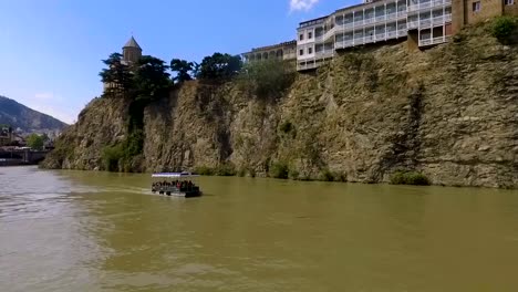 Touristen-auf-Boot-schwimmt-auf-Fluss-Kura,-Tiflis,-Metekhi-Kirche-im-Hintergrund