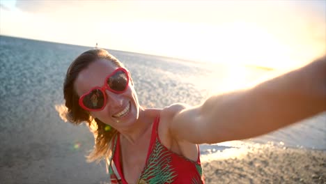 Junge-Frau-trägt-Herz-geformt-rote-Sonnenbrille-Selfie-Aufnahme-bei-Sonnenuntergang-am-schönen-Strand-von-Hawaii.-Slow-motion