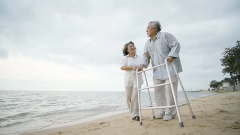 Senior-man-exercise-with-walker-and-his-wife-assist-him-walking-along-beach-in-slow-motion.-People-with-healthcare,-medical-and-retirement-concept.