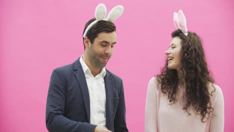 Young-beautiful-couple-standing-on-a-pink-background.-With-hackneyed-ears-on-the-head.-During-this-man-gives-his-wife-a-basket-of-colored-eggs.