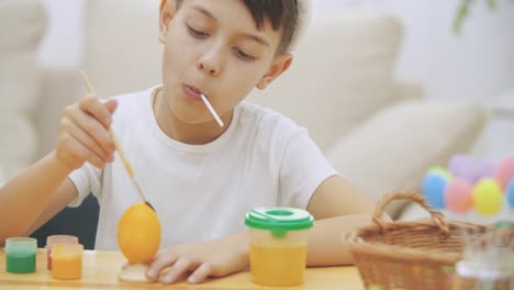 Intersted-and-concentrated-boy-is-finishing-to-colourize-an-Easter-egg-in-an-yellow-colour,-sitting-at-the-desk-and-tasting-sweet-lollipop.