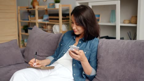 Kluge-Frau-schreibt-im-Notebook-mit-Smartphone-lächelnd-auf-Sofa-zu-Hause-sitzen