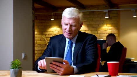 Closeup-shoot-of-old-caucasian-businessman-browsing-on-the-tablet-indoors-in-the-office-on-the-workplace