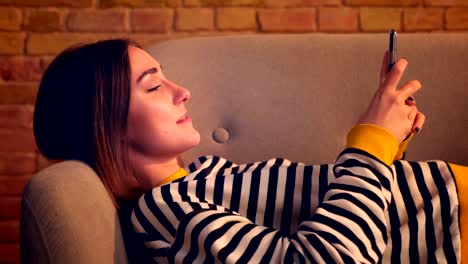 Primer-plano-vista-lateral-retrato-de-joven-chica-bonita-usando-el-teléfono-y-sonriendo-felizmente-acostado-en-el-sofá-en-un-acogedor-apartamento-en-el-interior