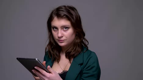 Closeup-shoot-of-adult-attractive-brunette-female-texting-on-the-tablet-and-looking-at-camera-with-background-isolated-on-gray