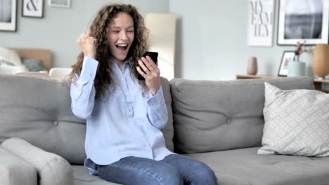 Lockly-Hair-Woman-jubelt-Erfolg-auf-Smartphone,-während-auf-der-Couch-sitzen