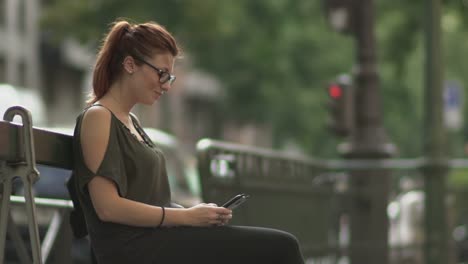 Cute-smiling-redhead-girl-with-glasses,-freckles,-piercings-and-red-hair-writing-a-text-message-on-her-smartphone-sitting-on-street-bench,-during-sunny-summer-in-Paris.-Slow-motion.-Trendy.