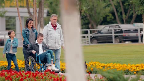 Happy-family-walks-on-the-road-with-his-son-in-a-wheelchair,-the-dog-runs-nearby.-Teenager-with-limited-opportunities-for-a-walk-with-his-parents,-sister-and-dog.