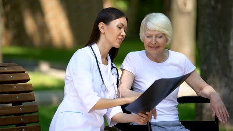 Smiling-nurse-showing-spinal-x-ray-to-old-woman-in-wheelchair-in-park,-recovery