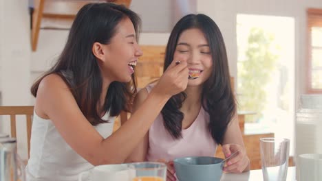 Asian-Lesbian-couple-has-breakfast-drink-juice,-cornflakes-cereal-and-milk-in-bowl-on-table-in-kitchen-in-the-morning.