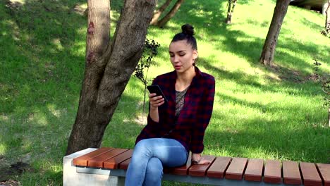 Young-beautiful-girl-in-a-plaid-shirt-is-sitting-on-a-park-bench,-smiling-and-writing-a-message-in-her-smartphone.