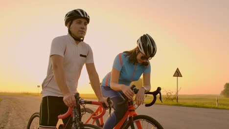 Un-hombre-y-una-mujer-con-cascos-con-bicicletas-de-pie-y-hablan-al-atardecer.-Descansar-después-de-un-paseo-en-bicicleta-en-la-carretera.-Bicicletas-de-pista.-Un-par-de-deportes.