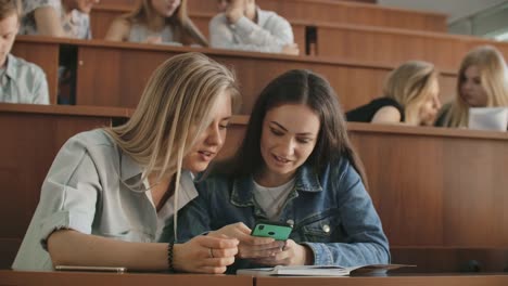 Chicas-bonitas-estudiantes-están-utilizando-el-teléfono-inteligente,-viendo-la-pantalla,-hablando-y-riendo-sentado-en-los-escritorios-de-la-universidad.-Redes-sociales,-internet,-millennials-y-concepto-educativo.