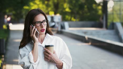 Dame-in-Brille,-weißes-Hemd.-Sie-spricht-auf-dem-Handy,-hält-Kaffee-in-Papiertasse,-eine-Pause-im-Park.-Geschäftskonzept.-Zeitlupe