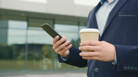 Un-hombre-de-negocios-se-encuentra-fuera-del-centro-de-negocios-con-una-taza-de-café-en-la-mano.-Está-enviando-mensajes-de-texto-en-su-teléfono-inteligente.-Lleva-traje-y-gafas.-Disparo-de-primer-plano.-4K