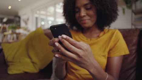 Shot-of-young-woman-sitting-on-sofa-holding-mobile-in-her-hand-checking-messages