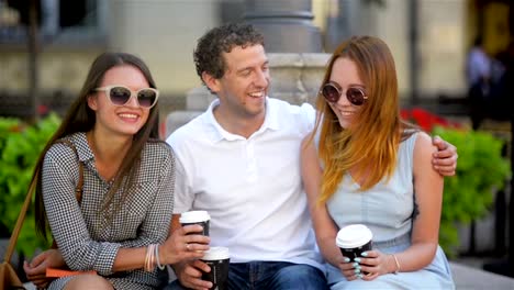 Porträt-von-drei-Freunden-Kaffeetrinken-sitzen-auf-der-Bank-im-Freien-in-der-Stadt-während-der-warmen-Sommertag.-Boy-ist-umarmt-Mädchen-im-blauen-Kleid-Licht.-Eine-andere-Frau-hat-lange-dunkle-Haare-und-Sonnenbrille