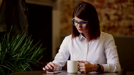 Filling-Brünette-mit-Brille-Nahaufnahme.-Getränke,-duftender-Kaffee-und-verwenden-ein-Mobiltelefon-wählt-sich-SMS-Nachrichten,-Nachrichten-Uhren,-blättert-durch-Fotos,-in-einem-sozialen-Netzwerk-kommuniziert