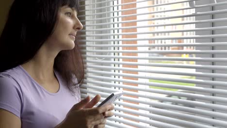 Mujer-joven-con-smartphone-por-ventana-en-página-de-inicio.-Primer-plano,-seria-niña-mirando-por-la-ventana-utiliza-un-smartphone.-Mensaje-de-texto,-navegar-por-Internet,-aplicación,-red-social.