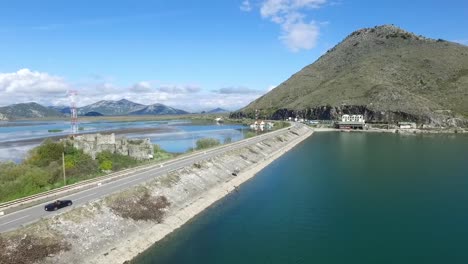 Coche-negro-en-fondo-de-mar,-montañas-y-cielo-de-puente-azul.-Material-de-archivo.-Coche-va-en-un-camino-cerca-del-río,-vista-aérea.-Material-de-archivo.-Vista-aérea-en-bosque-pequeño-lago-y-línea-de-carretera-pasando-junto-a-la