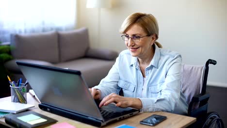 Exitosa-mujer-senior-en-silla-de-ruedas-trabajando-en-ordenador-portátil,-charlando-con-el-cliente