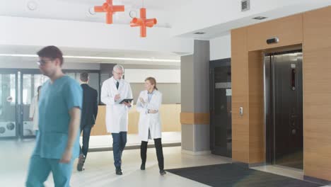 In-the-Hospital-Young-Couple-Walks-to-Reception.-Medical-Personnel,-Doctors,-Nurses,-Assistants-and-Patients-in-the-Busy-Lobby.-New,-Modern-Medical-Facility.