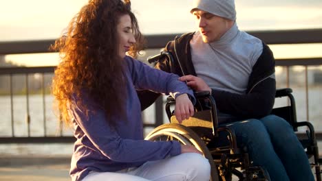 Happy-couple---disabled-man-in-wheelchair-talking-with-attractive-young-woman-enjoing-the-sunset