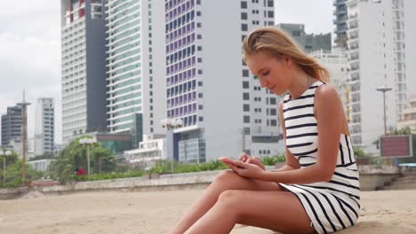 Junge-schöne-Frau,-schlank-mit-langen-blonden-Haaren-in-schwarzen-und-weißen-Kleid-ist-am-Strand-sitzen-und-per-Smartphone-auf-Hintergrund-der-Stadt.-Mädchen-am-Strand-durch-Berühren-des-Bildschirms.