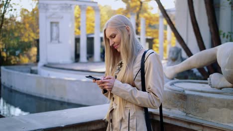 Mujer-rubia-alegre-en-la-capa-de-color-beige-en-el-parque-de-otoño,-mirando-su-smartphone