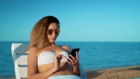 Young-Smiling-Mixed-Race-Tourist-Girl-in-Sonnenbrillen-Mit-Handy-am-Strand-in-der-Nähe-des-Meeres-mit-schönen-Sonnenuntergang