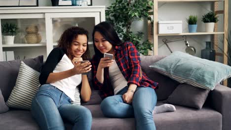 Fröhliche-Studenten-Asiaten-und-Afroamerikaner-reden-und-lachen-Blick-auf-Smartphone-Bildschirm-mit-Gadgets-sitzen-auf-dem-Sofa-zu-Hause.-Jugend-und-Gerätekonzept.