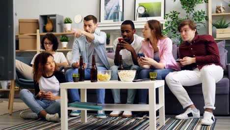 Cheerful-friends-using-smartphones-then-watching-TV-and-talking-discussing-news