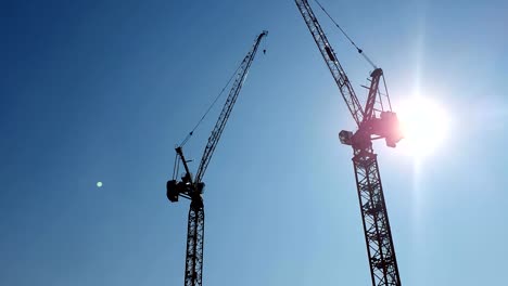 Dos-grúas-de-construcción-en-el-fondo-del-cielo-azul-y-el-sol,-que-brilla-en-la-cámara-que-se-esconde-detrás-de-la-grúa-de-construcción