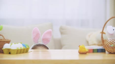 Linda-niña-con-orejas-de-conejo-se-esconde-debajo-de-la-mesa-llena-de-decoraciones-de-Pascua.-La-pequeña-chica-blanca-está-haciendo-caras.-Laught-está-en-la-habitación.