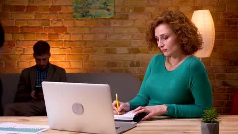 Closeup-shoot-of-adult-businesswoman-typing-on-the-laptop-taking-notes-indoors-in-the-office.-Female-and-male-employees-walking-on-the-background