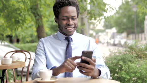 Hombre-de-negocios-africano-aclamación-éxito-en-Smartphone-sentado-en-al-aire-libre-Cafe