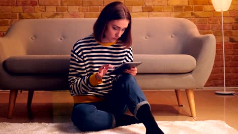 Closeup-portrait-of-young-pretty-girl-using-the-tablet-sitting-on-the-floor-in-a-cozy-apartment-indoors