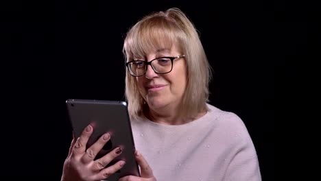 Closeup-shoot-of-old-caucasian-blonde-grandma-in-glasses-surfing-socail-media-on-the-tablet-in-front-of-the-camera-with-background-isolated-on-black