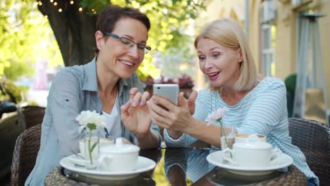 Aufgeregte-Frauen-mit-Smartphone-lachen-im-Open-Air-Café-in-der-Stadtstraße