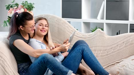 Zwei-glückliche-junge-charmante-lesbische-Mädchen-Blick-auf-Bildschirm-von-Tablet-PC-Umarmung-liegend-auf-der-Couch