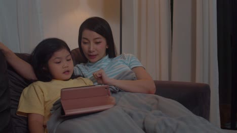 Joven-familia-asiática-e-hija-feliz-usando-la-tableta-en-casa.-Madre-coreana-relajarse-con-la-niña-viendo-películas-tumbadas-en-el-sofá-antes-de-ir-a-dormir-en-la-sala-de-estar-en-casa-en-la-noche.