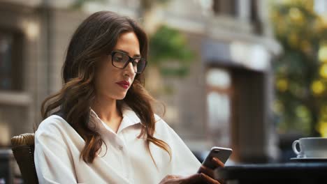 Geschäftsfrau-in-Brille,-weißes-Hemd.-Sitzen-am-Tisch-mit-Tasse-Kaffee-im-Café-im-Freien.-Nachrichten-auf-dem-Handy-durchsuchen,-sich-umsehen.-Zeitlupe