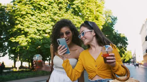 Chicas-sosteniendo-té-frío-en-vasos-de-plástico.-Riendo,-mirando-sorprendido-viendo-video-en-el-teléfono-móvil.-Caminando-por-la-avenida-de-la-ciudad.-De-cerca,-cámara-lenta