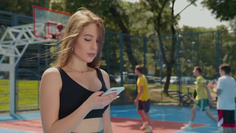 Ein-sportliches-Mädchen-textet-auf-ihrem-Smartphone.-Ihr-Haar-fliegt-im-Wind.-Sie-trägt-eine-Sportuniform.-Die-Leute-spielen-Floorball-im-Hintergrund.-4K