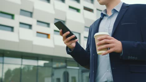 A-businessman-is-walking-near-the-business-center-with-a-cup-of-coffee-in-his-hand.-He-is-texting-on-a-smartphone.-He-is-wearing-a-suit-and-glasses.-Close-up-shooting.-4K