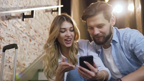 Vista-cercana-de-la-mujer-rubia-feliz-y-guapo-y-el-hombre-barba-que-usando-el-teléfono-mientras-está-sentado-en-los-escalones-dentro