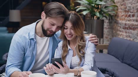 Fröhliche-moderne-stilvolle-junge-Paar-umarmt-und-beobachten-am-Telefon-der-Frau,-während-am-Tisch-in-der-Hotellobby-sitzen